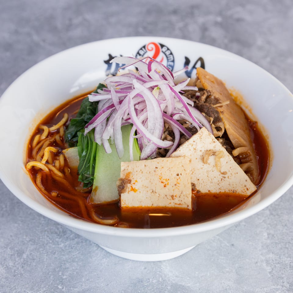 Spicy ramen bowl with noodles, bok choy, tofu, beef, bamboo shoots, and red onions.