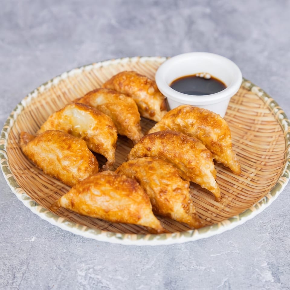 Golden-brown fried dumplings arranged on a woven plate with a small white bowl of soy sauce.