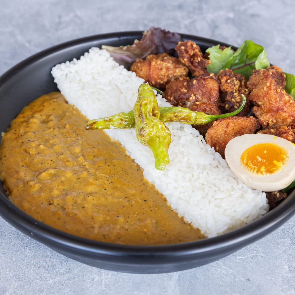 Japanese curry with rice, fried chicken, a soft-boiled egg, and green peppers in a black bowl.
